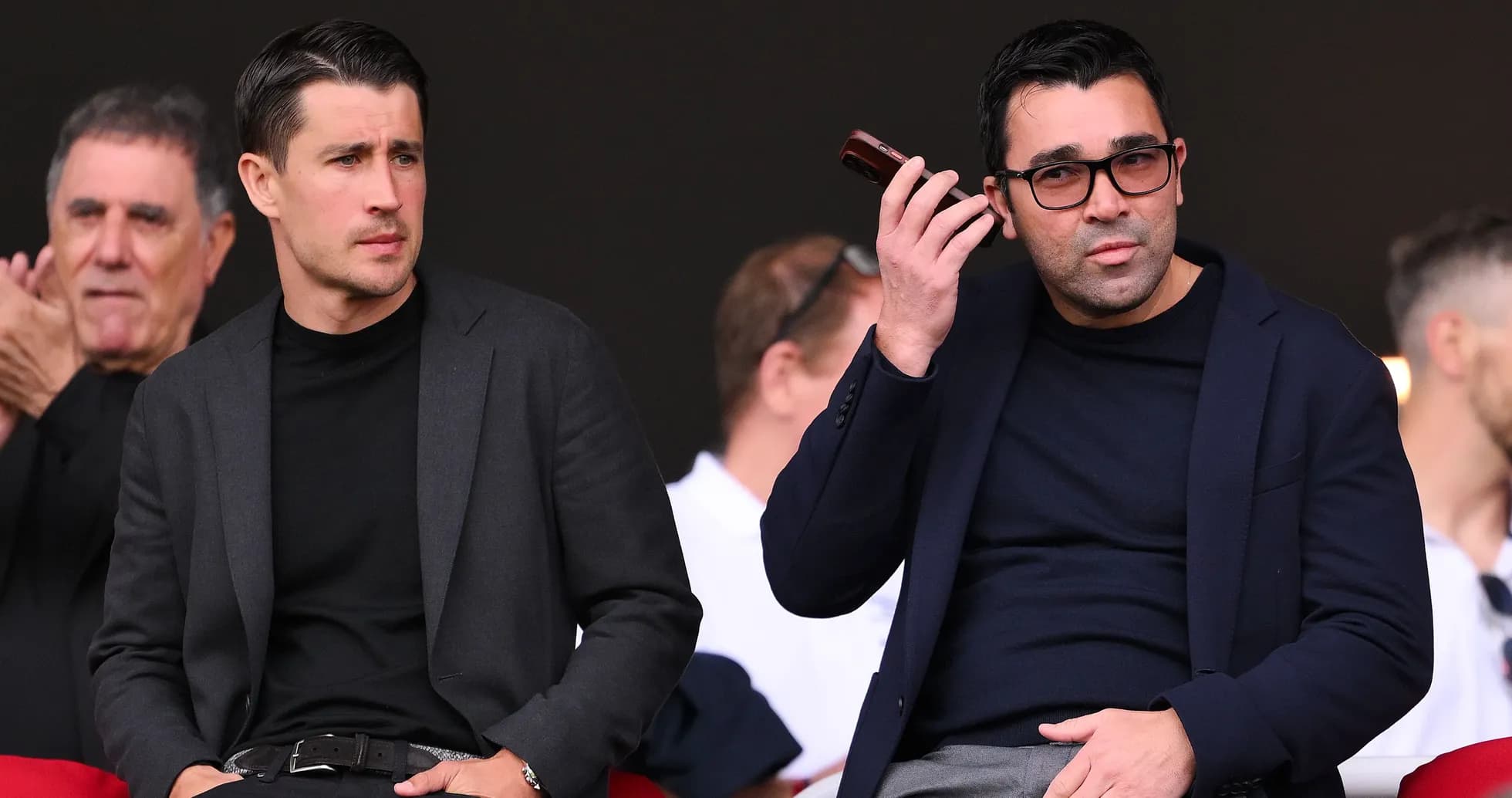 Two men seated in a stadium, one on phone Two men seated in a stadium, one on phone
