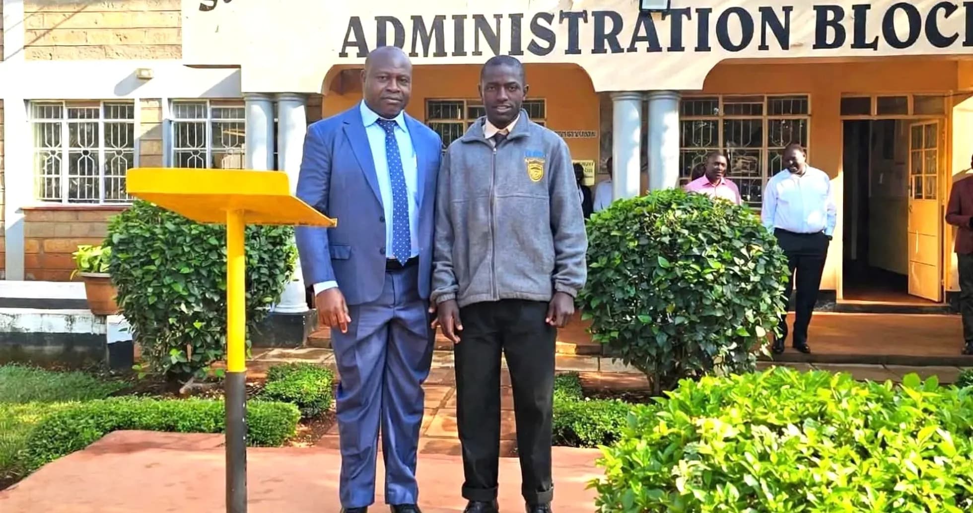 Two individuals posing in front of school building Two individuals posing in front of school building